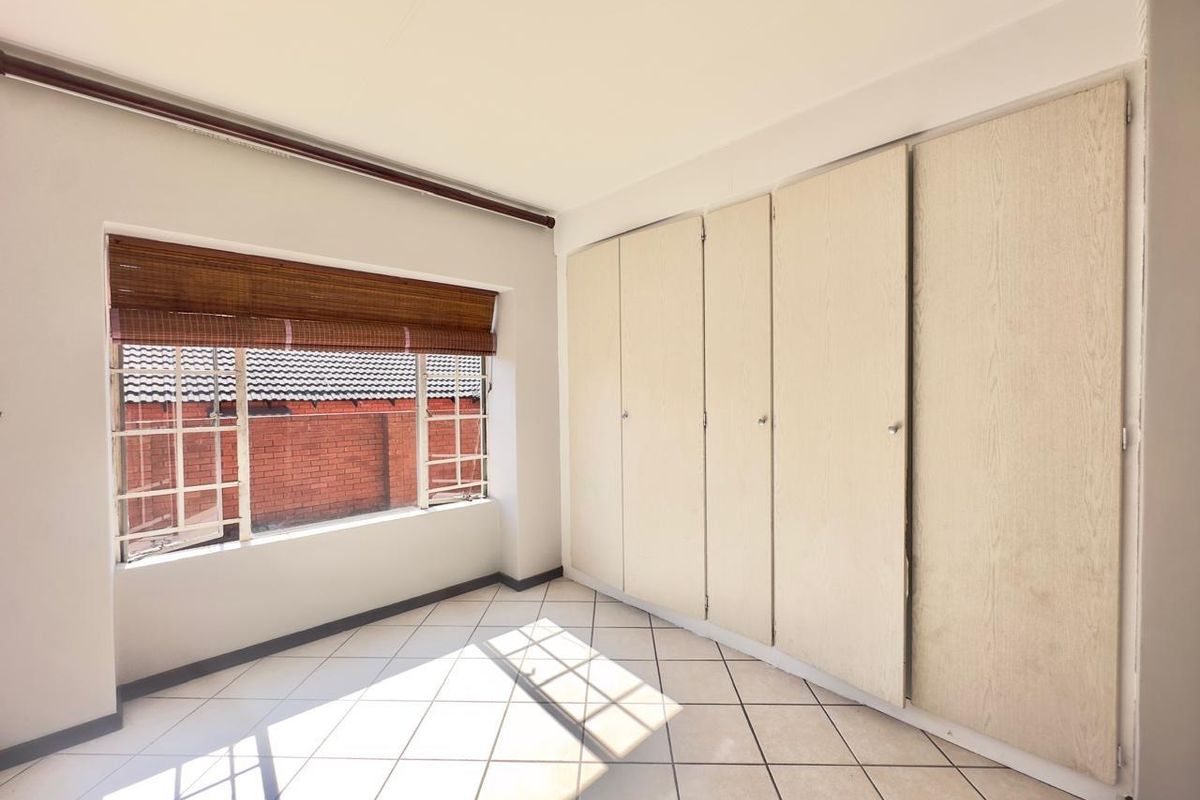 A bright, empty bedroom featuring tiled flooring, large built-in cupboards, and steel-framed windows with a view of the neighboring brick wall.