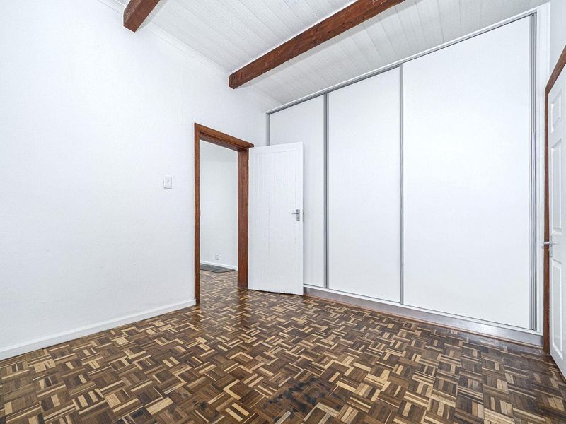 A bright, empty bedroom featuring classic parquet flooring, exposed timber ceiling beams, and large built-in cupboards with sliding doors.