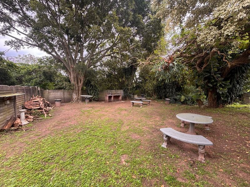 A spacious outdoor garden area featuring a built-in brick braai and mature trees, enclosed by a precast concrete boundary wall.