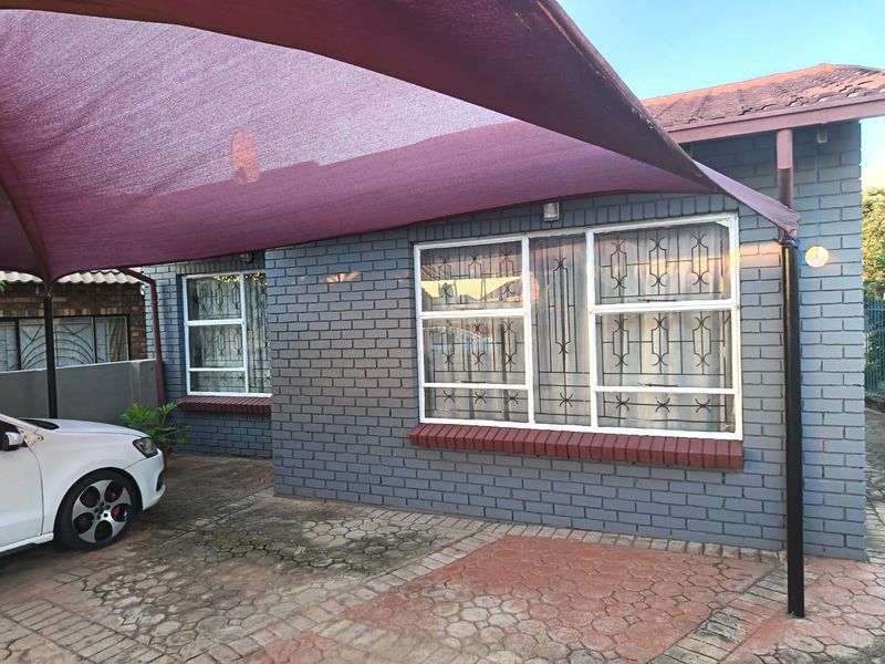 Front exterior view of a brick house featuring a paved driveway, security-barred windows, and a large shade sail covering the parking area.