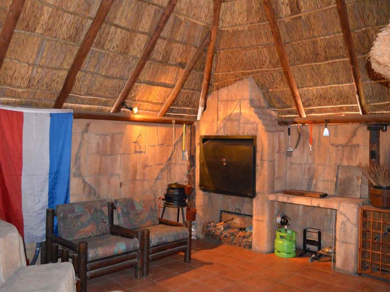 Rustic covered patio area featuring a traditional thatched roof, exposed timber rafters, and a built-in stone braai fireplace with terracotta flooring.