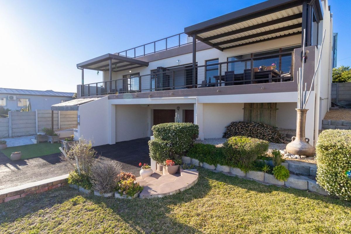 A rear view of a multi-level house featuring a spacious covered balcony, paved driveway, and landscaped garden area.