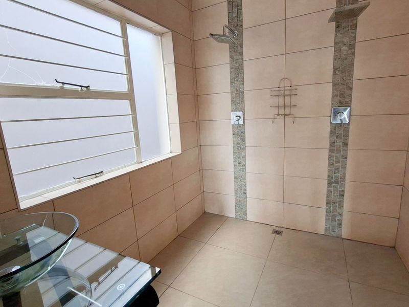 A spacious bathroom featuring a walk-in shower with dual shower heads, neutral wall and floor tiling, and a frosted window with security bars.