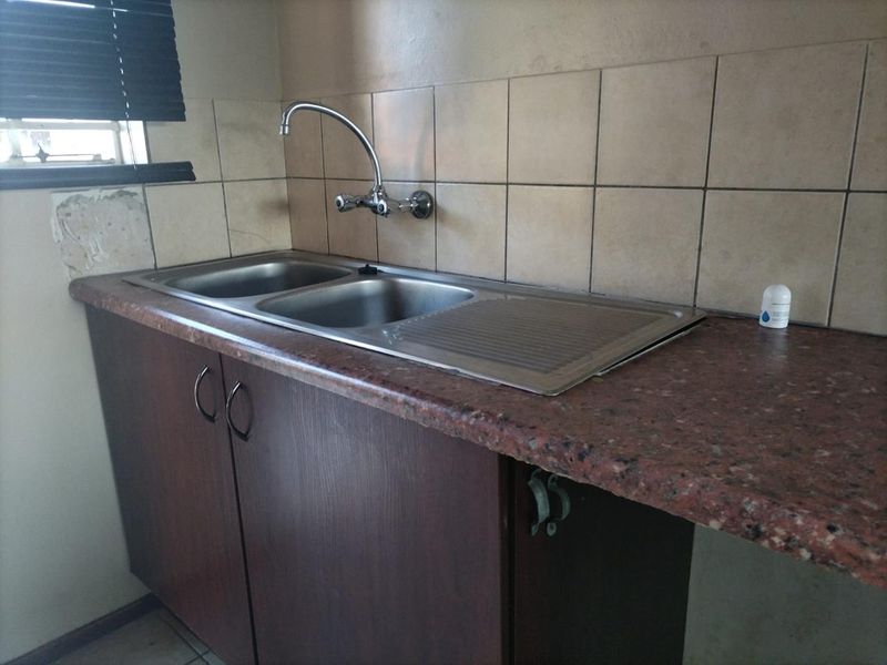 A functional kitchen area featuring a double bowl stainless steel sink, laminate countertops, and tiled splashbacks, requiring some cosmetic repairs to the wall and cabinetry.