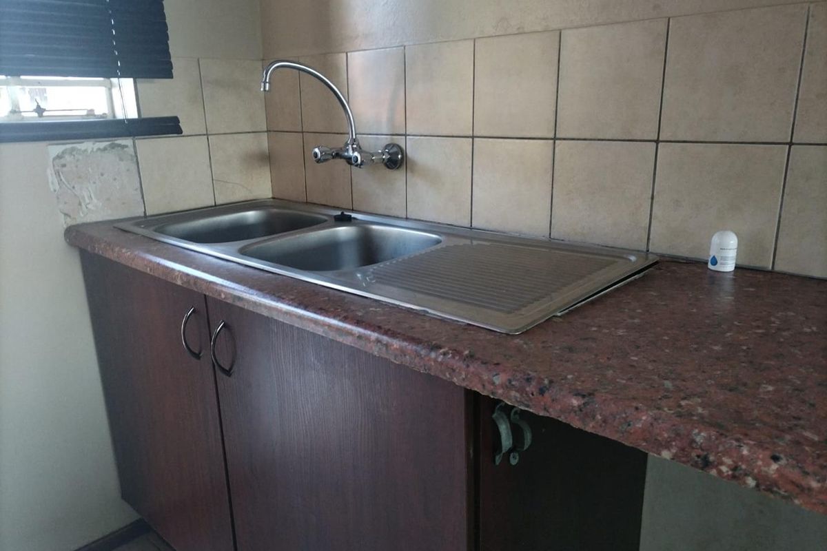 A functional kitchen area featuring a double bowl stainless steel sink, laminate countertops, and tiled splashbacks, requiring some cosmetic repairs to the wall and cabinetry.