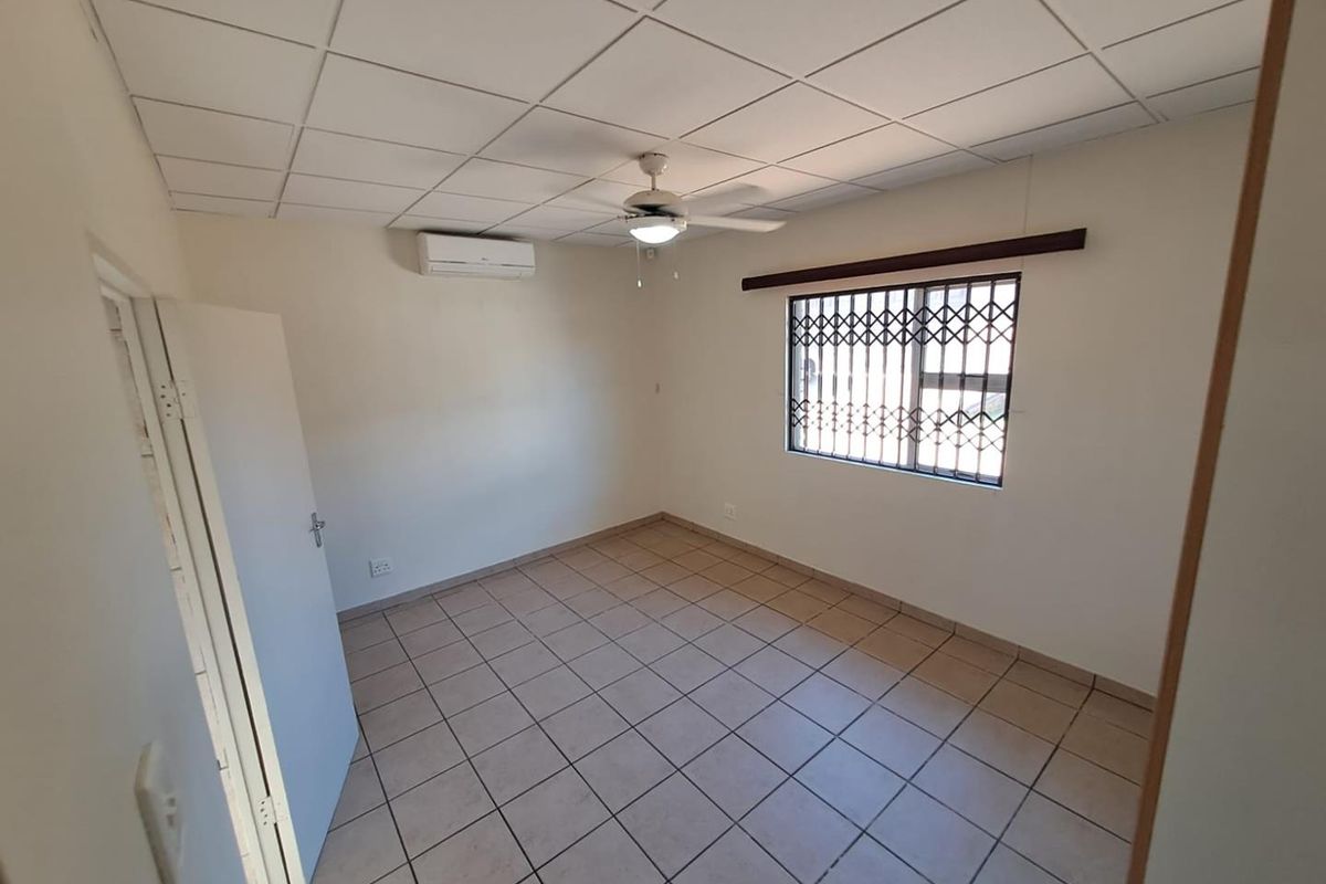 A clean, empty bedroom featuring tiled flooring, a drop ceiling, a ceiling fan, and a wall-mounted air conditioning unit with security bars on the window.