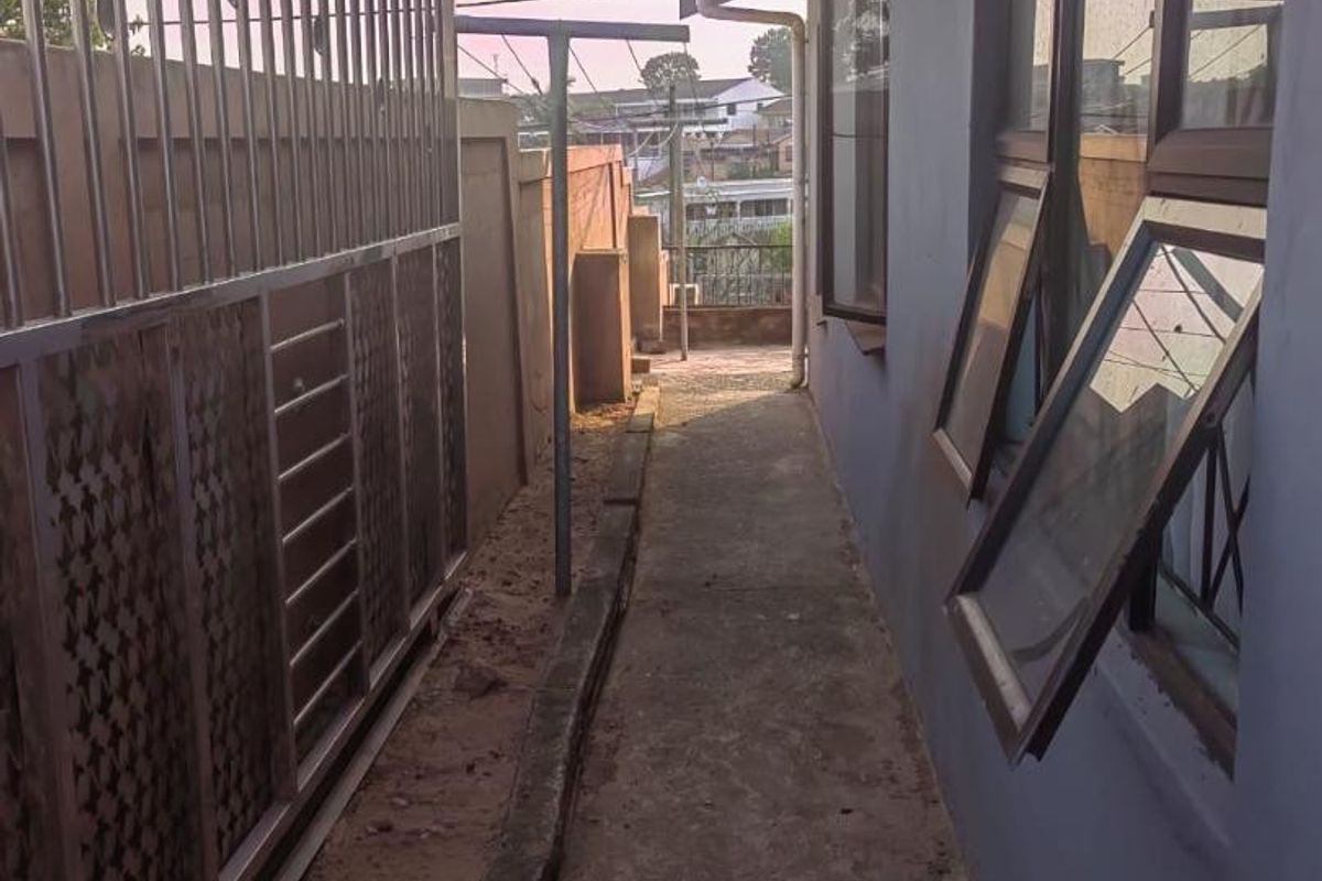 A narrow side exterior pathway featuring a metal security gate, concrete flooring, and casement windows along the side of the house.