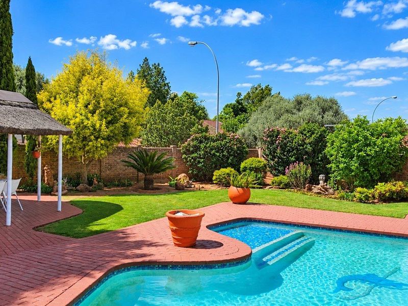 A well-maintained outdoor pool area featuring a swimming pool, brick-paved patio, and a thatched gazebo set within a landscaped garden.