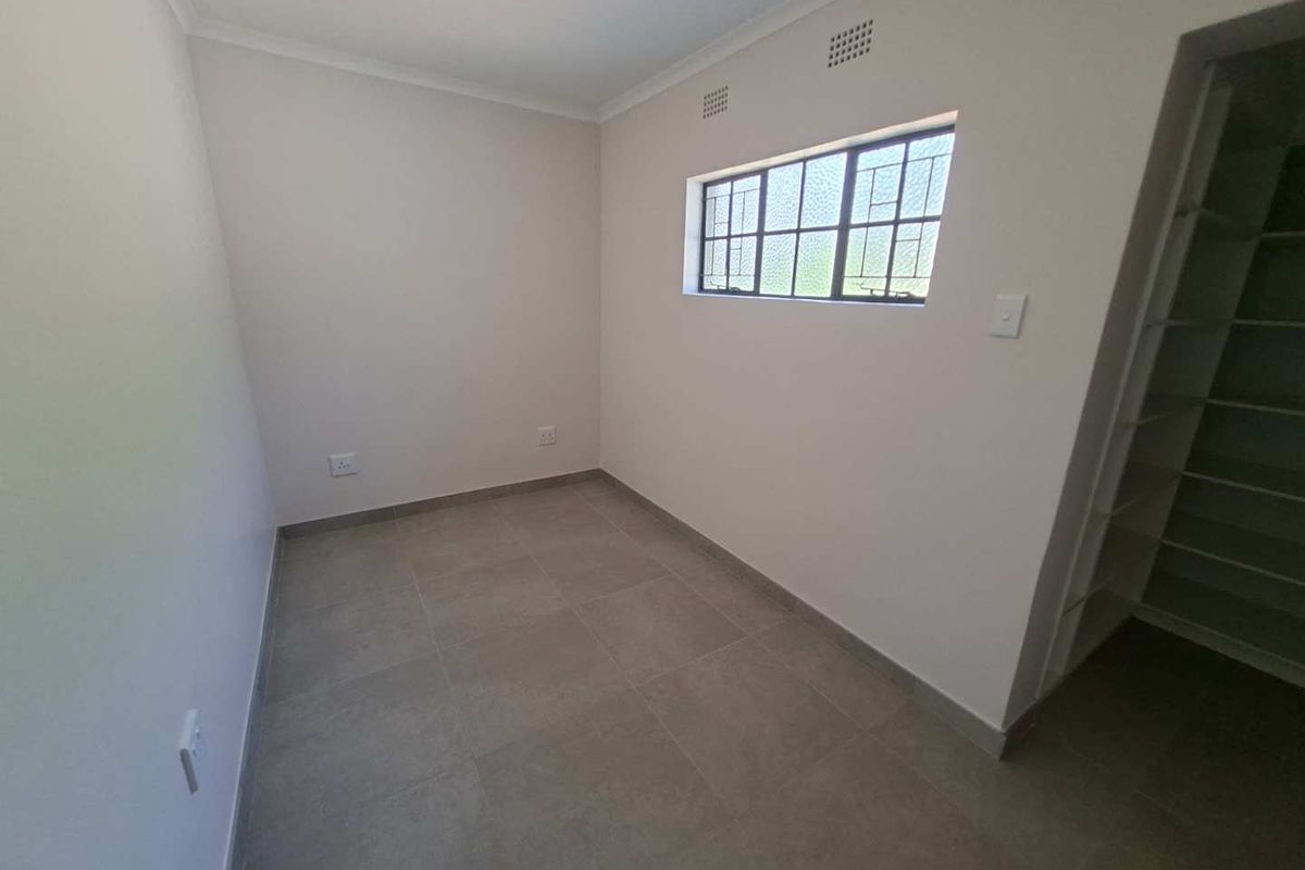 A clean, empty bedroom featuring neutral tiled flooring, built-in shelving, and a window with security bars.