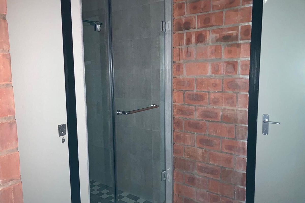 A bathroom shower enclosure featuring a glass door, tiled interior, and an exposed brick feature wall.
