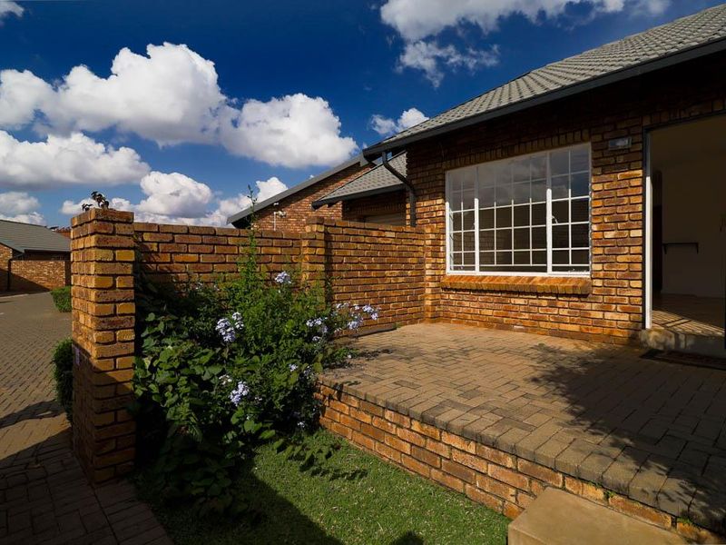 A well-maintained townhouse exterior featuring a face brick facade, a paved front patio, and a secure entrance with a metal gate.