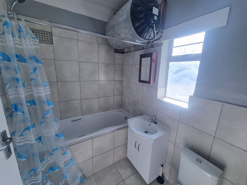 A functional bathroom featuring a bathtub with shower, vanity basin, and toilet, with tiled finishes and an exposed overhead geyser unit.
