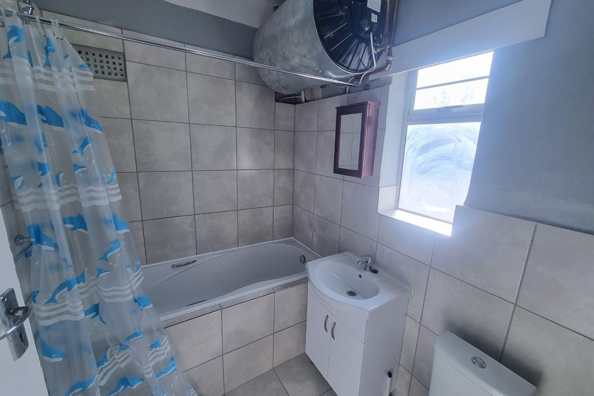 A functional bathroom featuring a bathtub with shower, vanity basin, and toilet, with tiled finishes and an exposed overhead geyser unit.