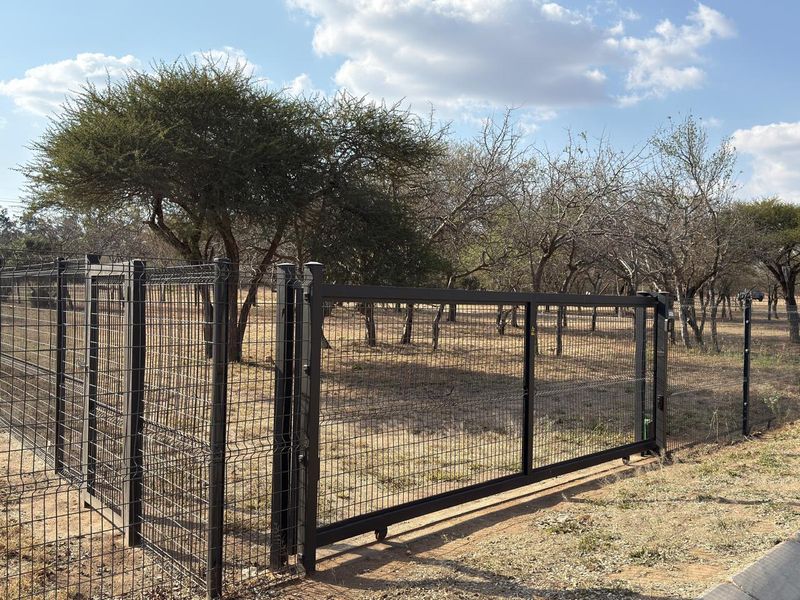 A secure metal mesh perimeter fence with a sliding gate entrance, enclosing a vacant plot with established trees.