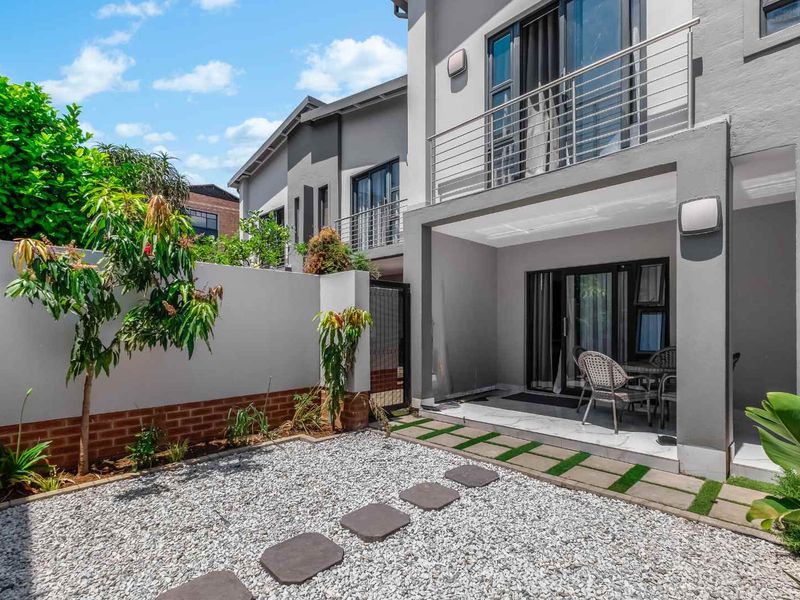 A well-maintained rear exterior featuring a covered patio, an upper-level balcony with stainless steel railings, and a landscaped gravel garden with stepping stones.