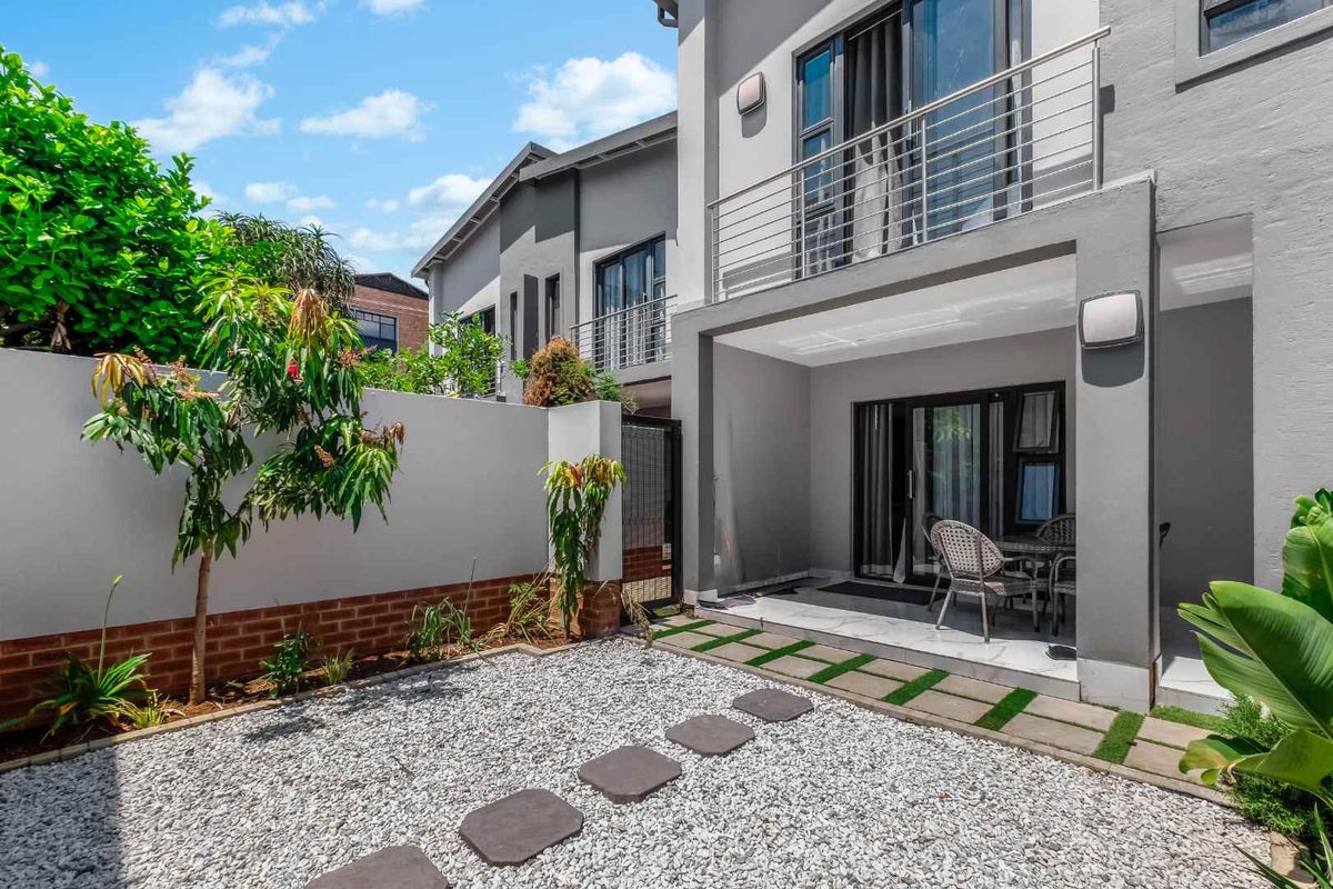 A well-maintained rear exterior featuring a covered patio, an upper-level balcony with stainless steel railings, and a landscaped gravel garden with stepping stones.