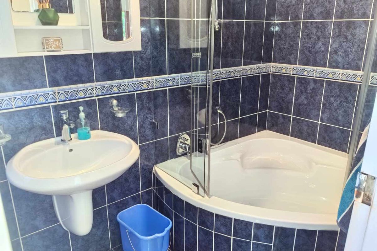 A bathroom featuring a corner bathtub with a glass shower screen, a pedestal sink, and dark blue wall tiling with decorative border accents.