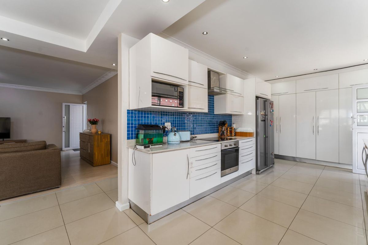 Modern kitchen featuring high-gloss white cabinetry, blue tiled backsplash, integrated oven, and sleek tiled flooring, seamlessly flowing into the adjacent living area.