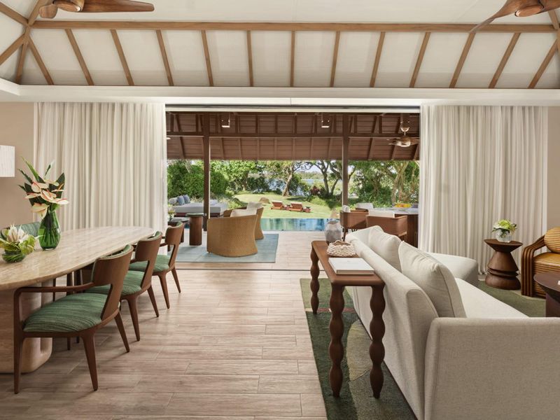 Spacious open-plan living and dining area featuring vaulted ceilings with exposed timber rafters, flowing seamlessly onto a covered patio overlooking the pool and garden.