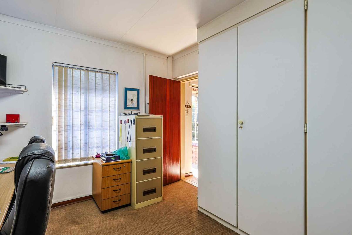 A functional bedroom or office space featuring built-in storage cupboards, carpeted flooring, and a window with vertical blinds.
