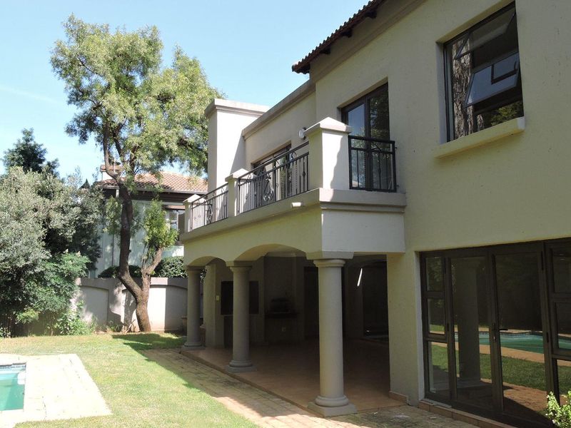 Rear view of the house featuring a covered patio, upper balcony with wrought iron railings, and a swimming pool set in a landscaped garden.