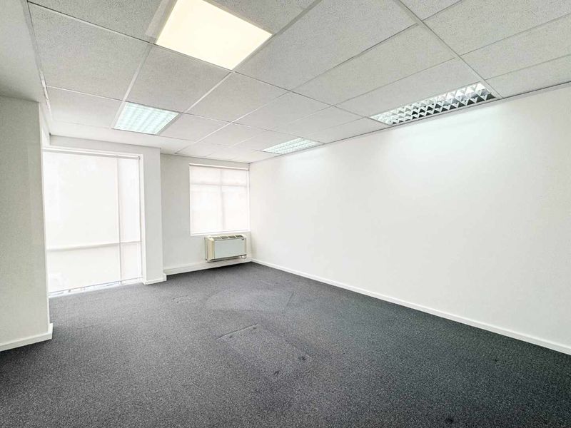 A bright, empty office space featuring a suspended ceiling with recessed lighting, carpeted flooring, and a wall-mounted air conditioning unit.
