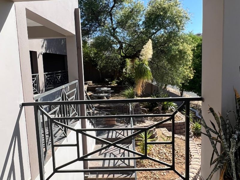 A covered balcony featuring a metal balustrade and tiled flooring, overlooking a landscaped garden area with retaining walls.