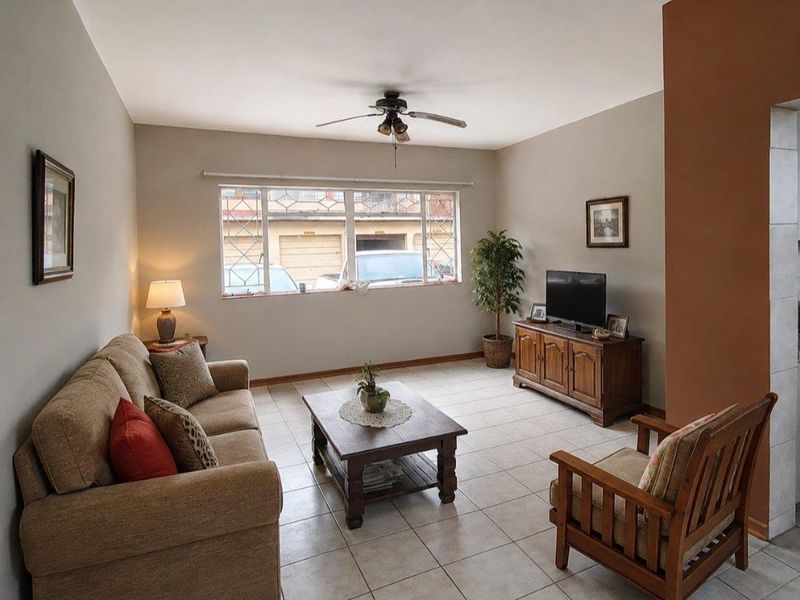 A well-lit living room featuring neutral tiled flooring, a ceiling fan, and large windows with security bars, offering a comfortable and functional living space.