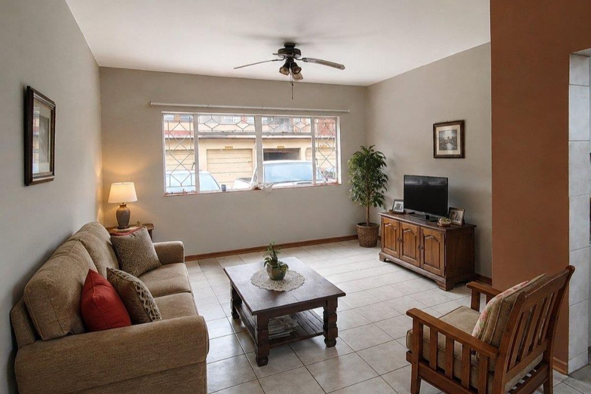 A well-lit living room featuring neutral tiled flooring, a ceiling fan, and large windows with security bars, offering a comfortable and functional living space.