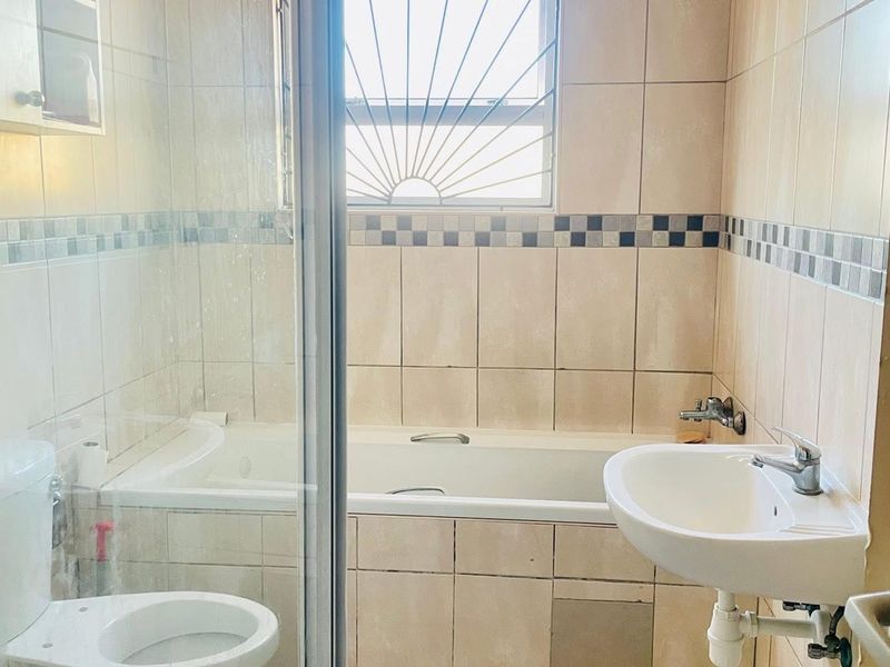 A functional bathroom featuring a built-in bathtub, glass shower enclosure, wall-mounted basin, and toilet with neutral wall tiling.