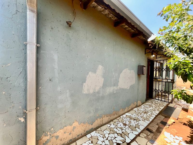 Rear exterior view showing a walkway with tiled finish, security gate, and wall surface requiring maintenance and repainting.