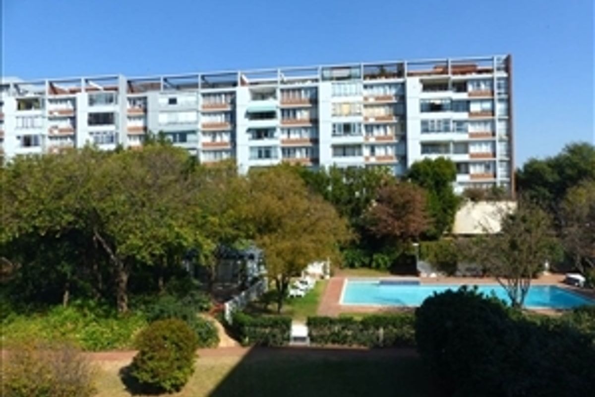 Exterior view of a multi-story apartment complex featuring a communal swimming pool, landscaped garden areas, and mature trees.