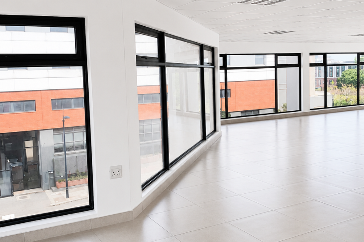 A bright, open-plan office space featuring large windows, neutral ceramic floor tiles, and a modern suspended ceiling with integrated lighting.