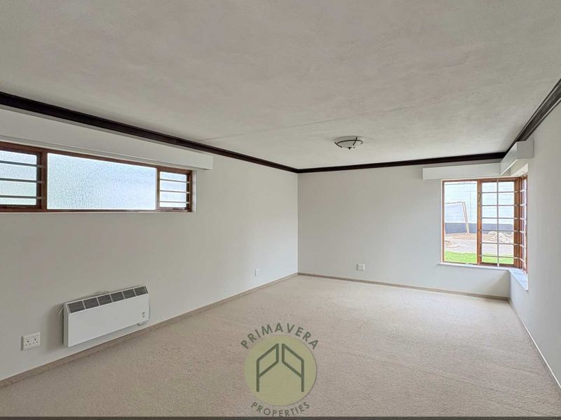 A spacious, empty living room featuring neutral carpeting, wall-mounted heating, and timber-framed windows with security bars.