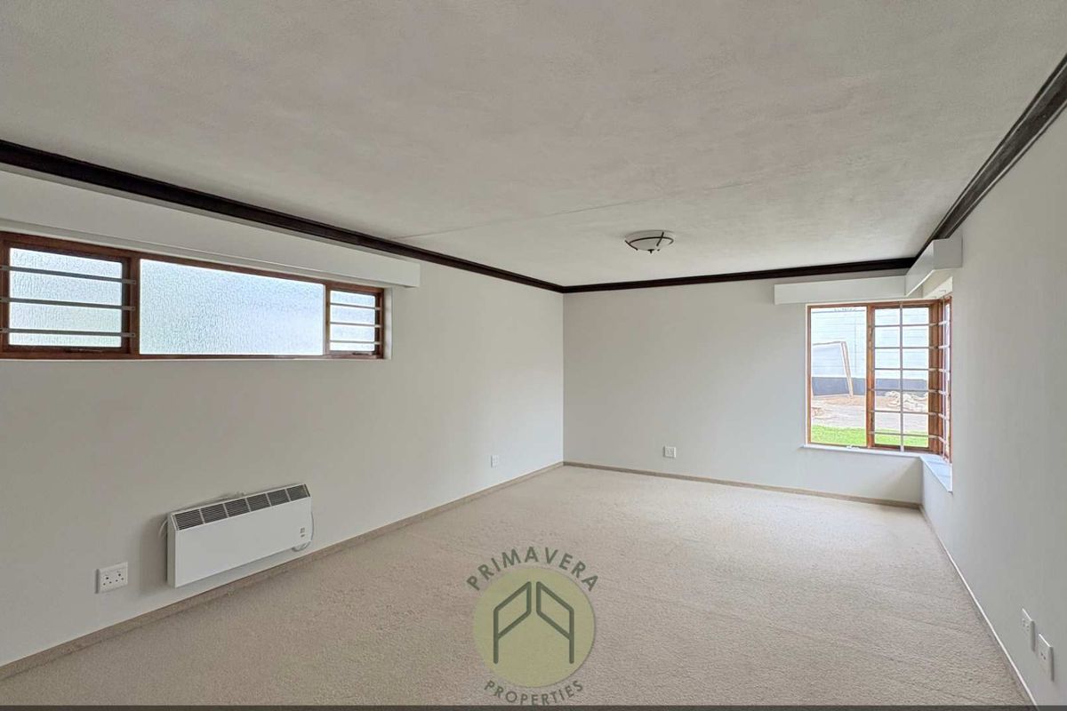 A spacious, empty living room featuring neutral carpeting, wall-mounted heating, and timber-framed windows with security bars.