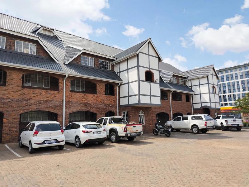 Exterior view of a multi-story apartment building featuring classic brickwork, Tudor-style architectural detailing, a metal roof, and an adjacent paved parking area.