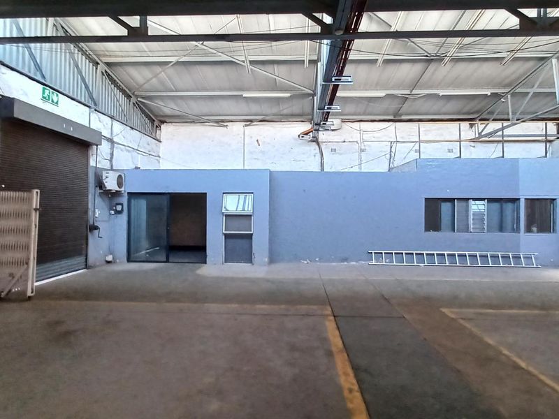 Spacious industrial warehouse unit featuring high ceilings with exposed steel trusses, concrete flooring, a roller shutter door, and an integrated internal office area.