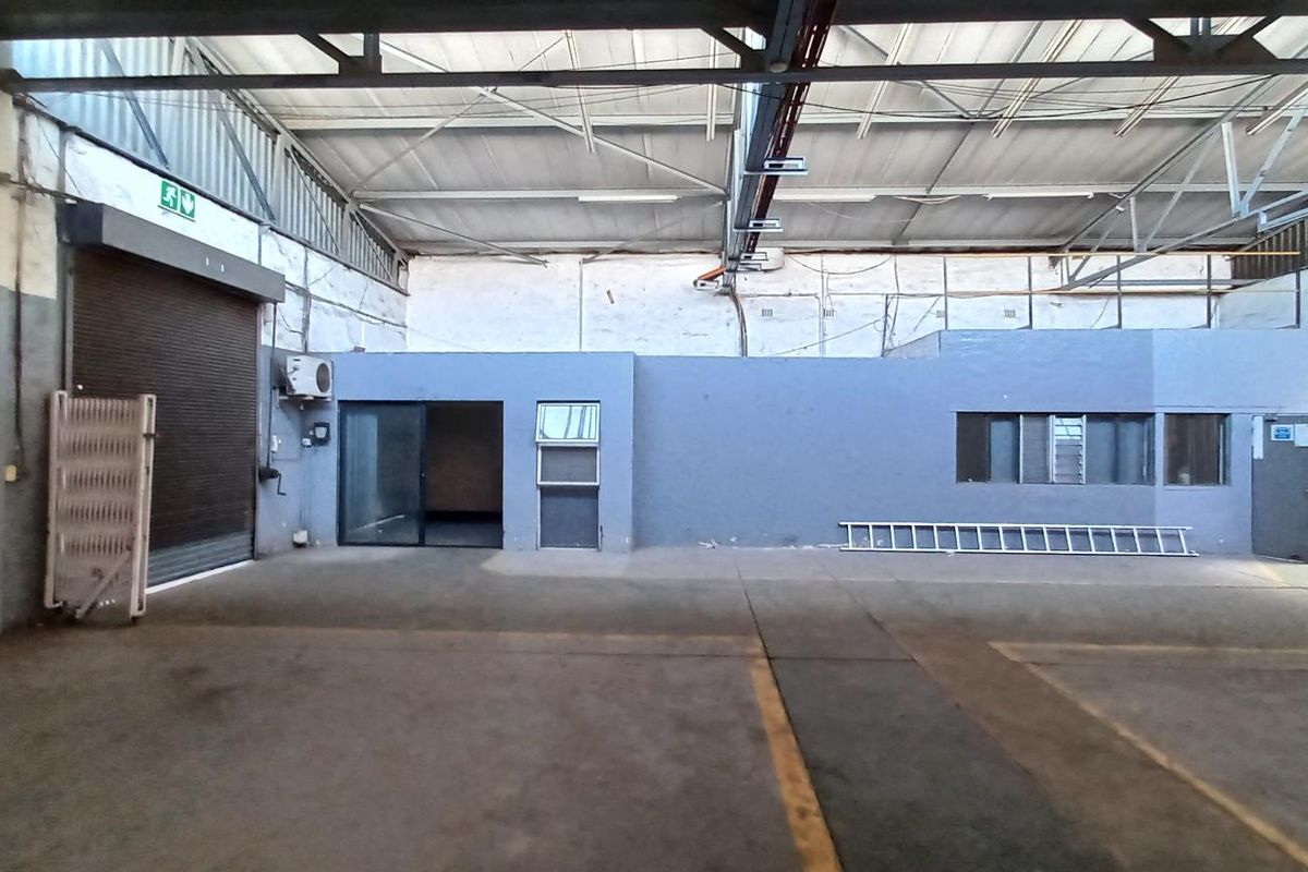 Spacious industrial warehouse unit featuring high ceilings with exposed steel trusses, concrete flooring, a roller shutter door, and an integrated internal office area.