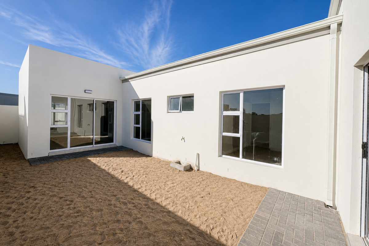 Rear exterior view of the house featuring modern white rendered walls, aluminium-framed windows, sliding glass doors, and a paved walkway leading to a sandy yard.