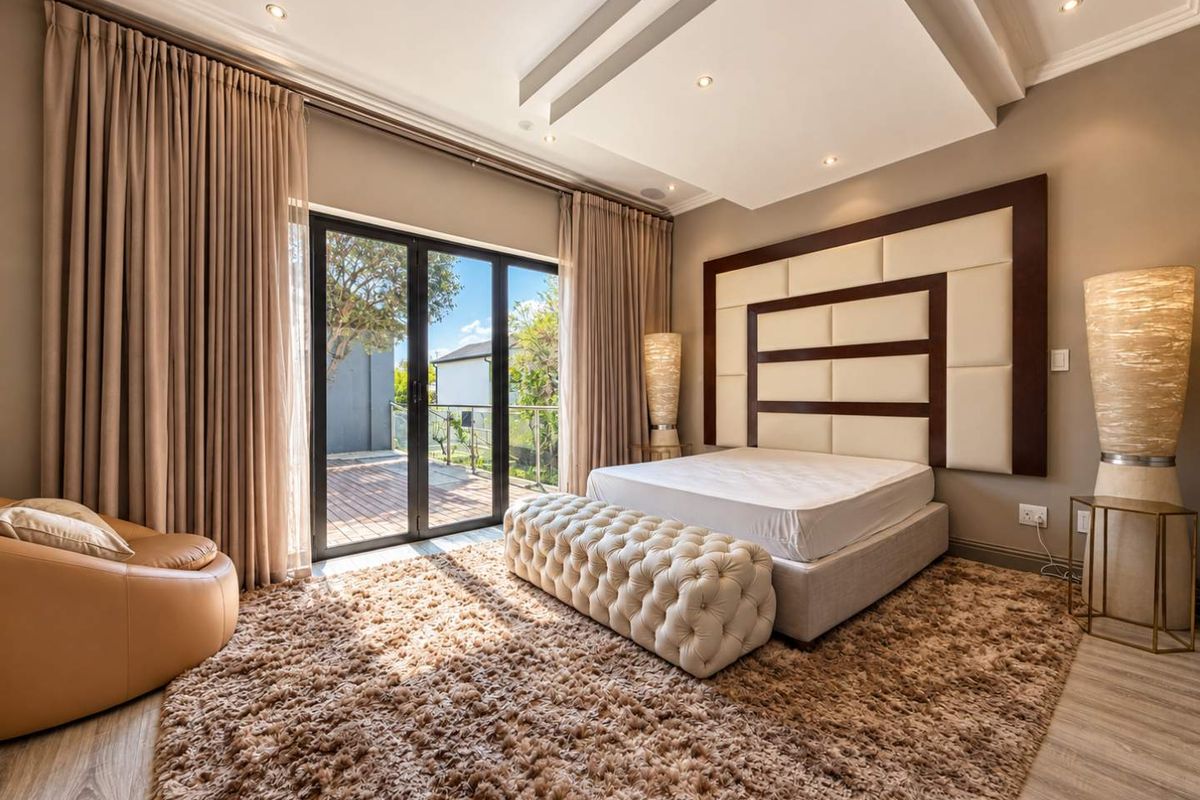 A spacious, well-lit bedroom featuring a recessed ceiling with downlights, a large upholstered headboard, and sliding glass doors leading to an outdoor deck.