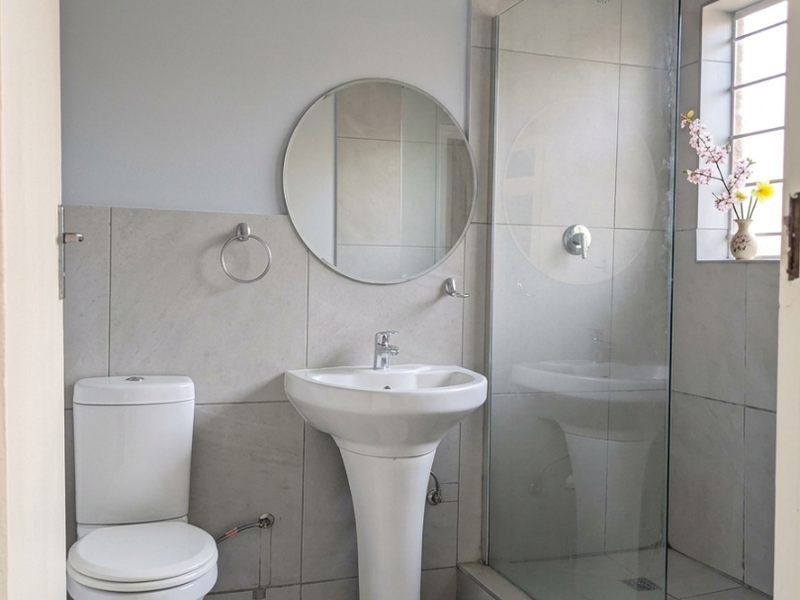 A modern, clean bathroom featuring a pedestal basin, toilet, and a glass-enclosed shower with neutral grey wall and floor tiles.