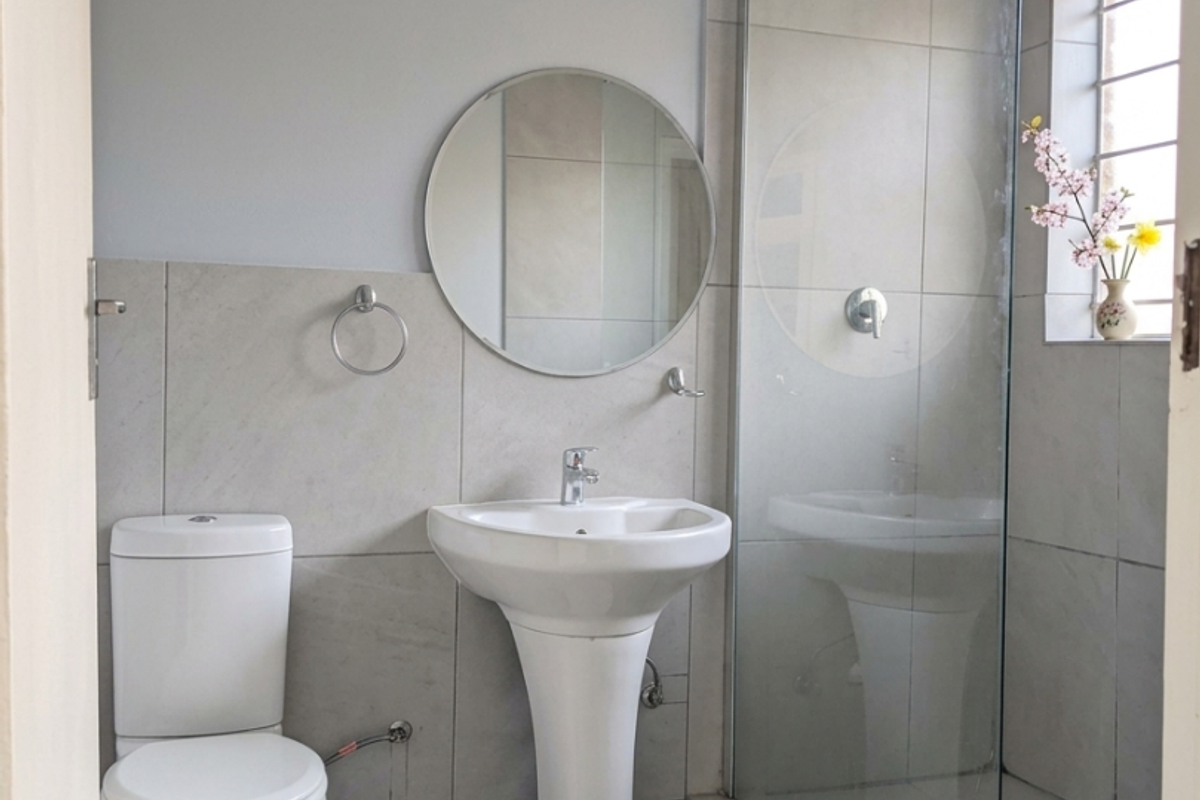A modern, clean bathroom featuring a pedestal basin, toilet, and a glass-enclosed shower with neutral grey wall and floor tiles.