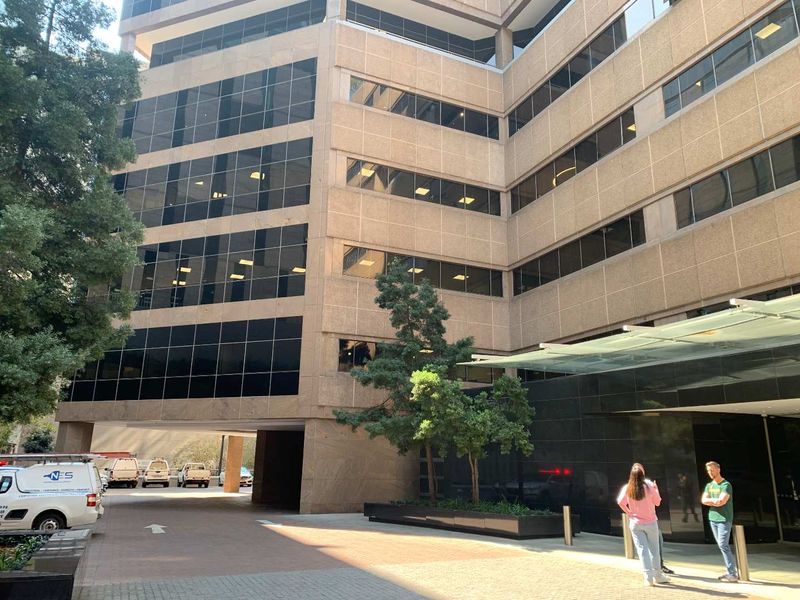 Exterior view of a multi-story commercial office building featuring a glass curtain wall facade, a modern glass entrance canopy, and a paved driveway area.
