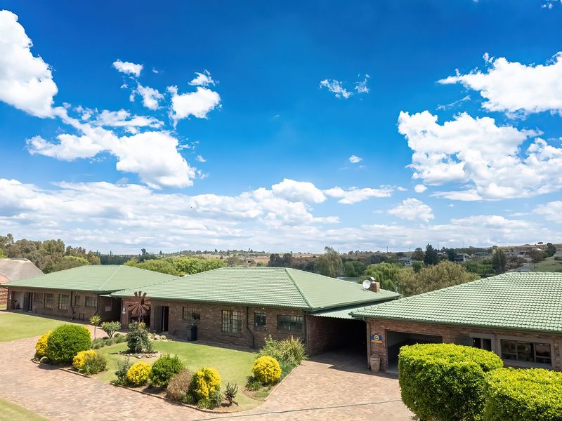 An elevated aerial view of a spacious brick house with a green tiled roof, featuring a large paved driveway and well-maintained garden.
