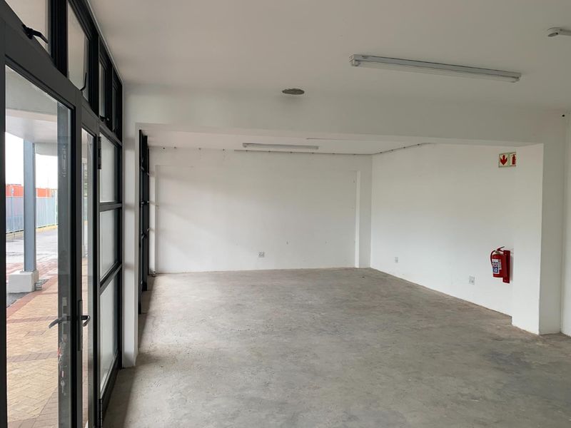 An empty, open-plan commercial space featuring black aluminium-framed glass doors, concrete flooring, and functional strip lighting, suitable for various business uses.