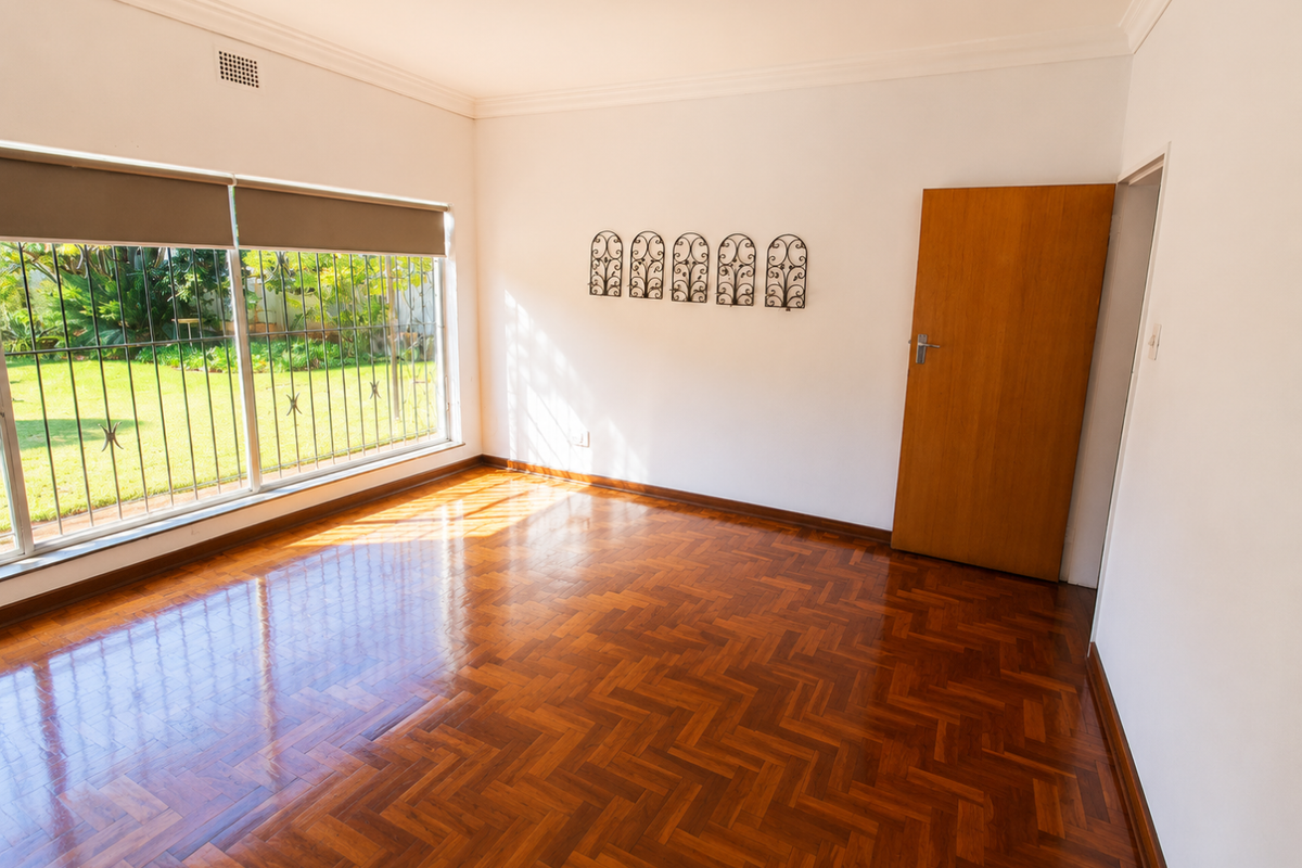 A bright, empty bedroom featuring polished herringbone parquet flooring, large windows with security bars, and neutral walls.