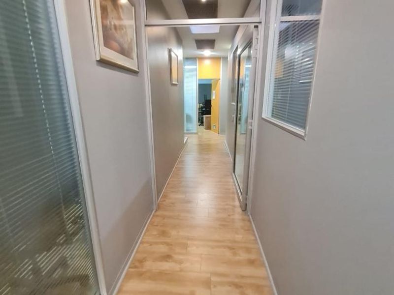 A clean, modern office hallway featuring wood-look laminate flooring, glass partition walls, and recessed ceiling lighting.