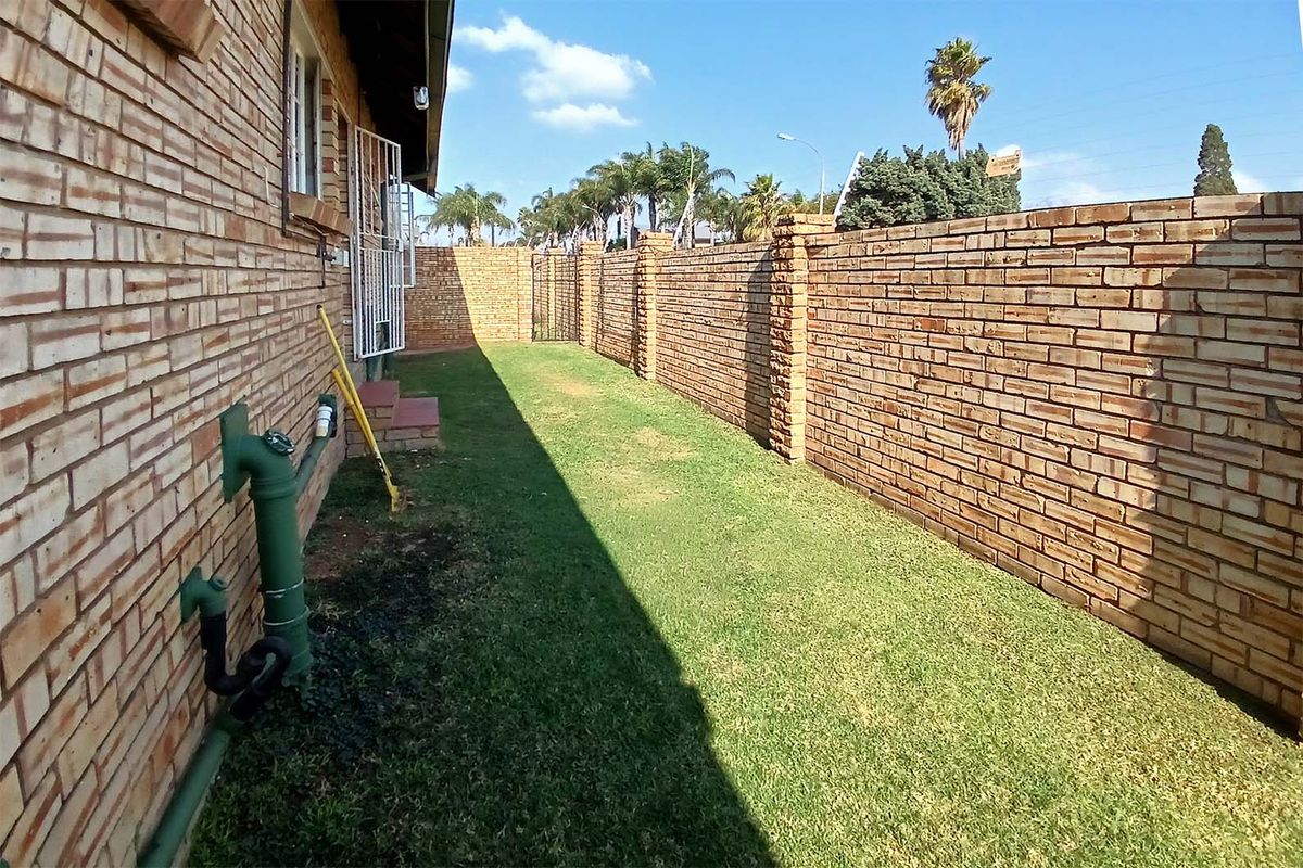 A side garden area featuring a face brick exterior wall, a long boundary wall, and established lawn.