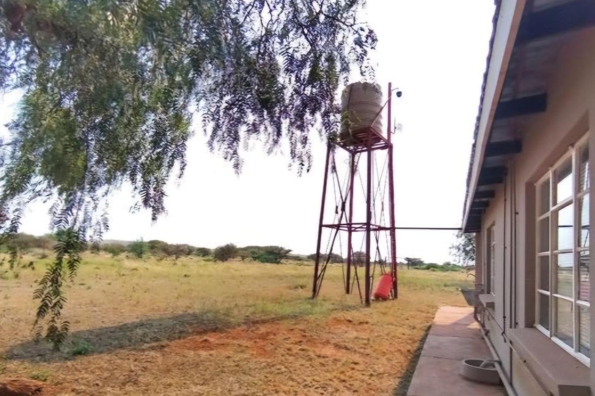 Rear exterior view of the property featuring a raised water tank on a steel stand, concrete walkway, and surrounding open farm landscape.