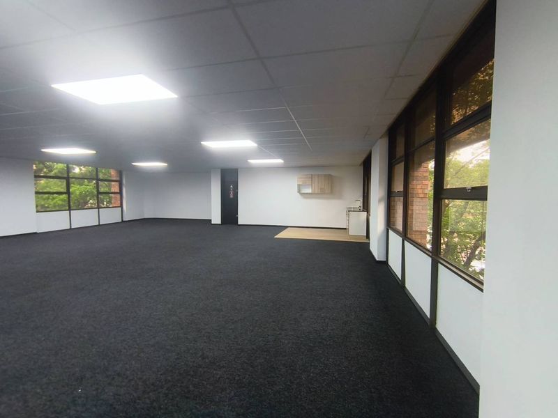 A spacious, open-plan office area featuring large windows, carpeted flooring, a suspended ceiling with recessed lighting, and a small kitchenette corner.